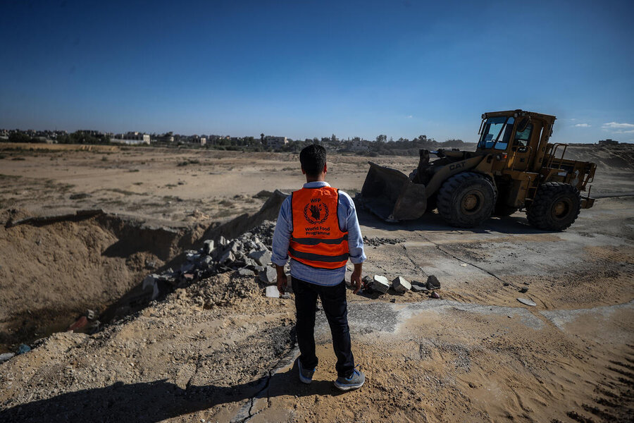 A man in an organ vest with a WFP logo on its back observes a construction tractor scooping rocks on a desert road