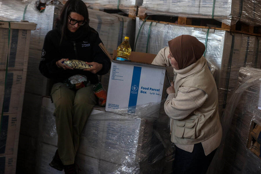 Two women check food items from a World Food Programme (WFP) food parcel in a warehouse filled with similar boxes. One is seated, examining a package, while the other unpacks.